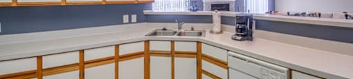 The clubhouse kitchen has a large breakfast bar that opens to the remainder of the clubhouse. The kitchen includes linoleum flooring, white cabinets with tan trim, a double basis sink, a dishwasher and coffee pot.