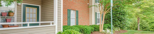 The apartment buildings are brick with tan siding and green shutters. The patios and balconies are enclosed. The building is surrounded by mature trees and lush landscape.