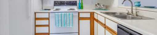 The three bedroom kitchen has white cabinets with tan trim, white appliances, formica countertops, and a double basin sink with gooseneck faucet. The breakfast bar has a place setting of fiesta ware for two.