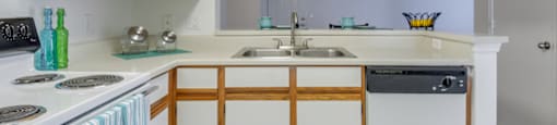 The three bedroom kitchen has white cabinets with tan trim, white appliances, formica countertops, and a double basin sink with gooseneck faucet. The breakfast bar has a place setting of fiesta ware for two.