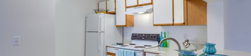 The three bedroom kitchen has white cabinets with tan trim, white appliances, formica countertops, and a double basin sink with gooseneck faucet. The breakfast bar has a place setting of fiesta ware for two.