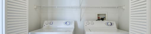 The laundry room is enclosed by two accordion, white, wooden doors. There is a washer and dryer with built-in shelving above them.