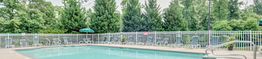 The community pool is rectangular in shape with blue and green pool furniture placed around the sundeck. There is a round picnic table with a green umbrella in the middle. The pool is placed on the back side of the Leasing Office.