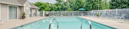 The community pool is rectangular in shape with blue and green pool furniture placed around the sundeck. There is a round picnic table with a green umbrella in the middle. The pool is placed on the back side of the Leasing Office.