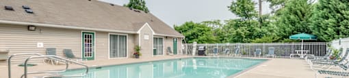 The community pool is rectangular in shape with blue and green pool furniture placed around the sundeck. There is a round picnic table with a green umbrella in the middle. The pool is placed on the back side of the Leasing Office.