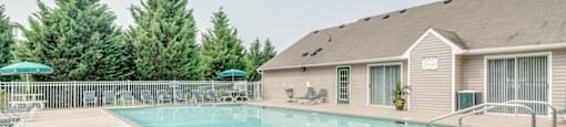 The community pool is rectangular in shape with blue and green pool furniture placed around the sundeck. There is a round picnic table with a green umbrella in the middle. The pool is placed on the back side of the Leasing Office.