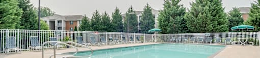 The community pool is rectangular in shape with blue and green pool furniture placed around the sundeck. There is a round picnic table with a green umbrella in the middle. The pool is placed on the back side of the Leasing Office.