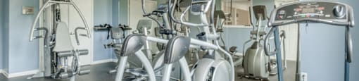 The community fitness center is equipped with a treadmill, two ellipticals and a weightlifting machine. One walls is made of mirrors and there is a TV mounted in the corner.