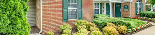 The Leasing Office building is mainly brick with a small amount of gray siding and forest green shutters and front door. There are flower beds on either side of the sidewalk leading to the front door.