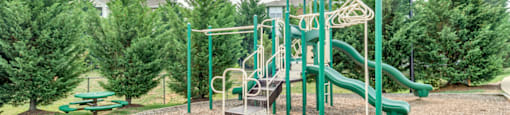 The community playground is surrounded by mulch and mature trees, and is located to the side of the clubhouse. The jungle gym is green and tan, has stairs leading to three slides and monkey bars.
