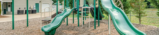 The community playground is surrounded by mulch and mature trees, and is located to the side of the clubhouse. The jungle gym is green and tan, has stairs leading to three slides and monkey bars.