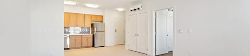 A spacious, empty room with white walls and a kitchen area in the background.