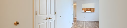A white door with a gold handle is on the left of a hallway with a wooden floor and a white wall.