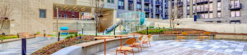 A wooden deck with chairs and a slide in the middle of a courtyard surrounded by buildings.