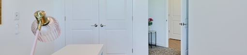 Now leasing senior 1-bedrooms in Baltimore, MD. A white table with a gold lamp stands in a room with white walls and a carpeted floor.