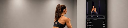 A woman is doing squats in a gym with a digital screen showing a workout video.