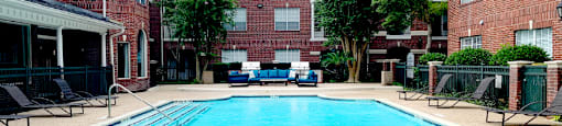 Luxury apartments with red brick exteriors, large swimming pool with spacious sundeck, grill, beautiful landscaping, lush landscaping, chaste trees, and pool-side lounge seating at Tuscany Apartments in Houston.
