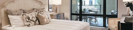 A carpeted bedroom with a king-size plush bed and a dresser overlooking a patio through a glass floor to ceiling window