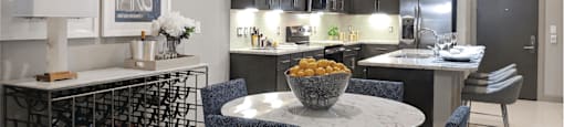 A round dining table with four chairs, a long wine racks along a wall and a view of an open kitchen with dark wood cabinetry