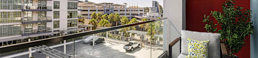 A balcony with a chair and a potted plant overlooks a parking lot and buildings.