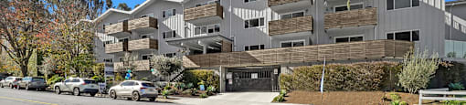 A modern building with a flat roof and balconies is situated on a street with cars parked in front.