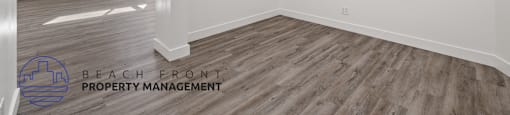 A spacious room with wooden flooring and white walls, with a logo for Beach Front Property Management in the bottom left corner.