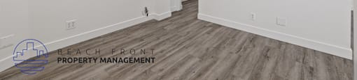 A room with wooden flooring and white walls, with a logo for Beach Front Property Management in the bottom left corner.