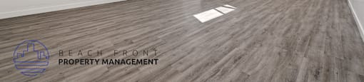 A spacious room with wooden flooring and white walls, with a logo for Beach Front Property Management in the bottom left corner.
