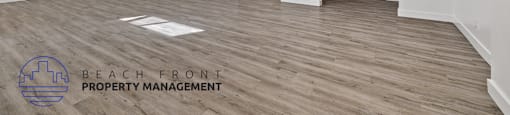 A spacious room with wooden flooring and white walls, with a logo for Beach Front Property Management in the bottom left corner.