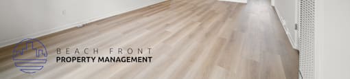 A spacious room with wooden flooring and white walls, with a logo for Beach Front Property Management in the bottom left corner.