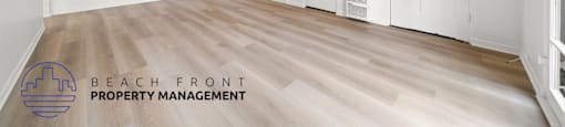 A spacious room with wooden flooring and white walls, with a logo for Beach Front Property Management in the bottom left corner.