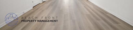 A spacious room with wooden floors and white walls, with a logo for Beach Front Property Management in the bottom left corner.