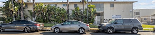 Three cars are parked in a lot with a building and palm trees in the background.
