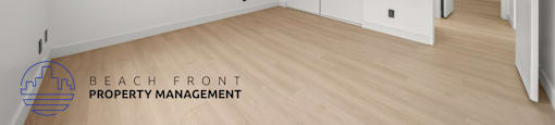 A room with white walls and wooden flooring, with a logo for Beach Front Property Management in the bottom left corner.