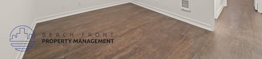 A spacious room with wooden flooring and white walls, with a logo for Beach Front Property Management in the bottom left corner.