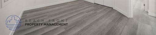 A spacious room with wooden flooring and white walls, with a logo for Beach Front Property Management in the bottom left corner.