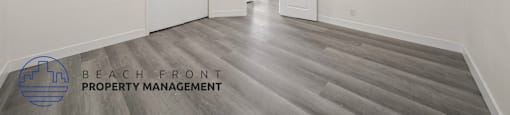 A spacious room with wooden flooring and white walls, with a logo for Beach Front Property Management in the bottom left corner.