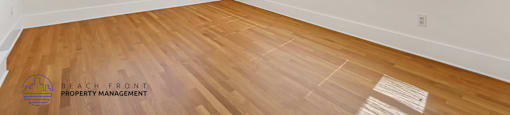 A room with wooden floors and white walls, with a window showing a view of a house.