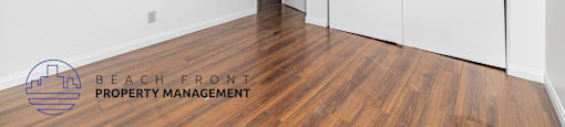 A room with a wooden floor and white walls, with a logo for Beach Front Property Management in the bottom left corner.