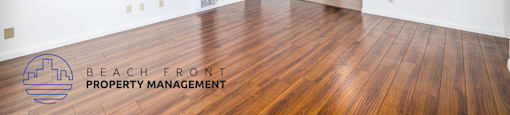 A spacious room with wooden flooring and white walls, with a logo for Beach Front Property Management in the bottom left corner.