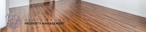A spacious room with wooden flooring and white walls, with a logo for Beach Front Property Management in the bottom left corner.
