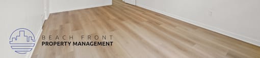 A spacious room with wooden floors and white walls, with a logo for Beach Front Property Management in the bottom left corner.