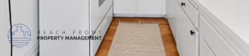 A kitchen with white appliances and cabinets, a rug on the floor, and a logo for Beach Front Property Management in the corner.