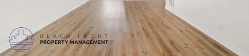 A spacious room with wooden floors and white walls, with a logo for Beach Front Property Management in the bottom left corner.