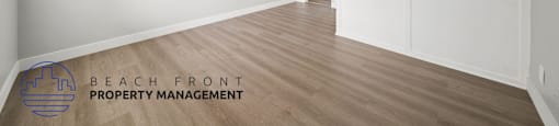 A spacious room with wooden flooring and white walls, with a logo for Beach Front Property Management in the bottom left corner.