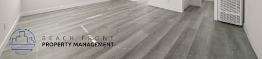A spacious room with wooden flooring and white walls, with a logo for Beach Front Property Management in the bottom left corner.