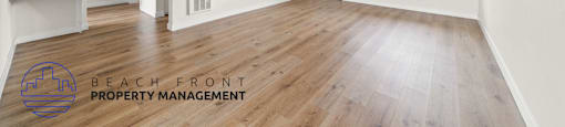 A spacious room with wooden flooring and white walls, with a logo for Beach Front Property Management in the bottom left corner.