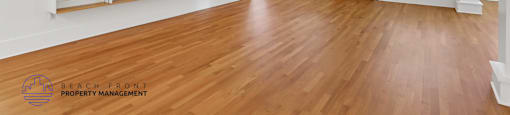 A spacious room with wooden floors and white walls, featuring a large window with blinds and a wooden door.