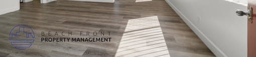 A spacious room with wooden flooring and white walls, with a logo for Beach Front Property Management in the bottom left corner.