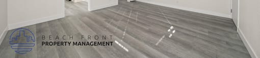 A spacious room with wooden flooring and white walls, with a logo for Beach Front Property Management in the bottom left corner.